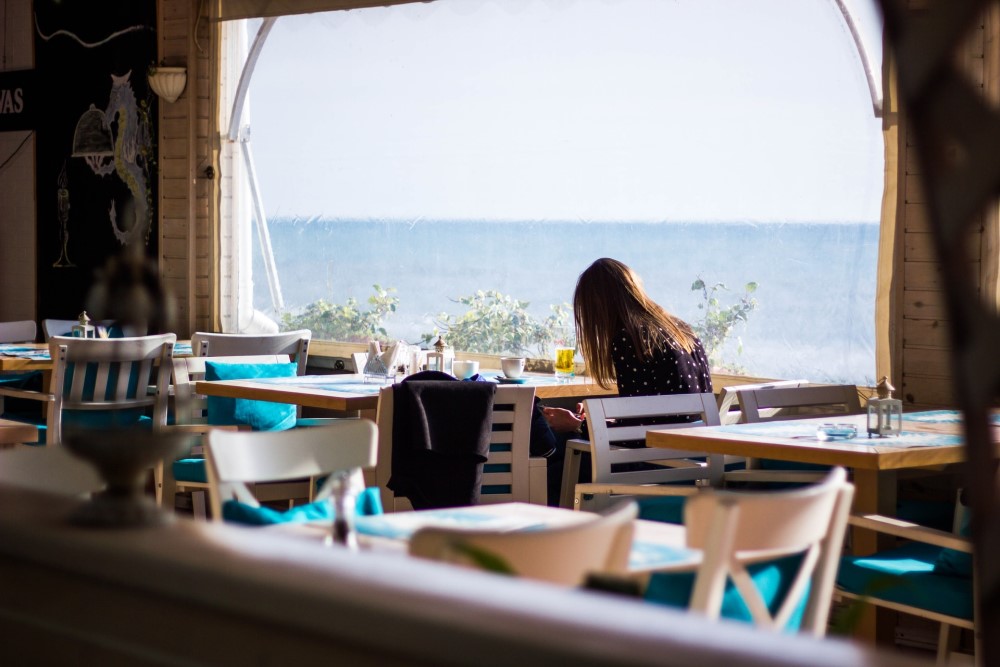 woman in cafe or restaurant looking at phone with sea beyond window